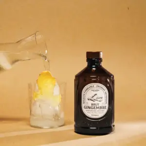 Isvann helles i et glass med fersk ingefær ved siden av en flaske Bacanha Brut Gingembre på beige bakgrunn.