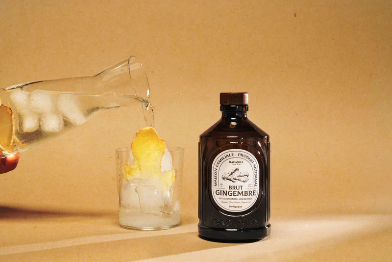 Isvann helles i et glass med fersk ingefær ved siden av en flaske Bacanha Brut Gingembre på beige bakgrunn.
