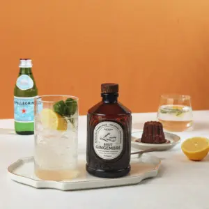 Glass med ingefærdrink og sitron står ved siden av en flaske Bacanha Brut Gingembre på et lyst bord med bakgrunn i varme toner.