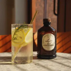 En flaske Bacanha Brut Gingembre og et glass med sitron og urter står på et steinbord i varmt sollys.