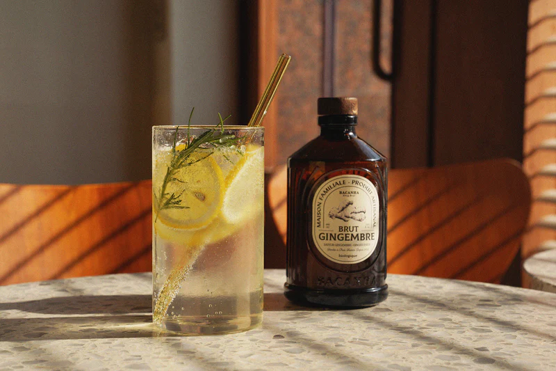 En flaske Bacanha Brut Gingembre og et glass med sitron og urter står på et steinbord i varmt sollys.