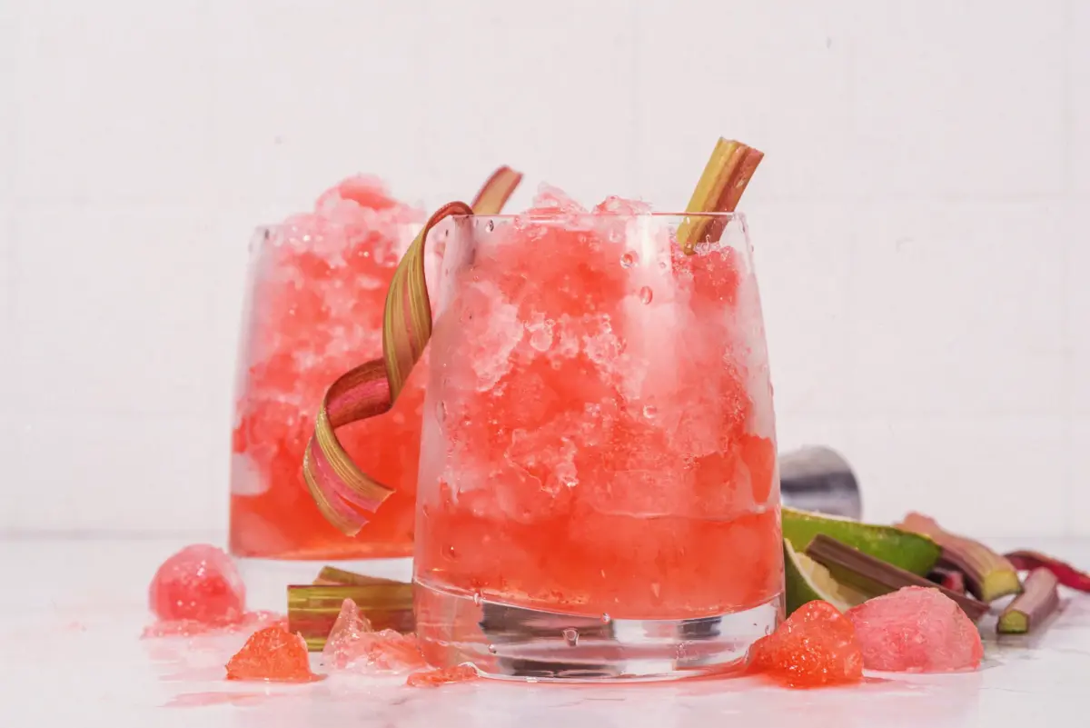 To glass med rosa bringebærdrink med is og rabarbraskall, servert på lys bakgrunn med friske ingredienser.