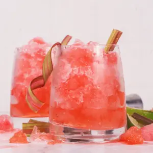 To glass med rosa bringebærdrink med is og rabarbraskall, servert på lys bakgrunn med friske ingredienser.