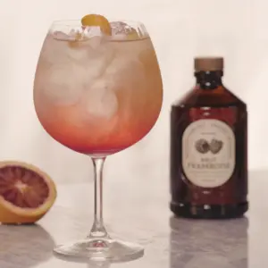 Et glass med bringebærdrink og is står foran en flaske Bacanha Brut Framboise, med sitrus i bakgrunnen.
