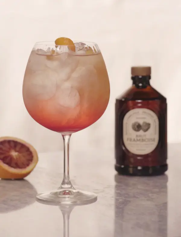 Et glass med bringebærdrink og is står foran en flaske Bacanha Brut Framboise, med sitrus i bakgrunnen.