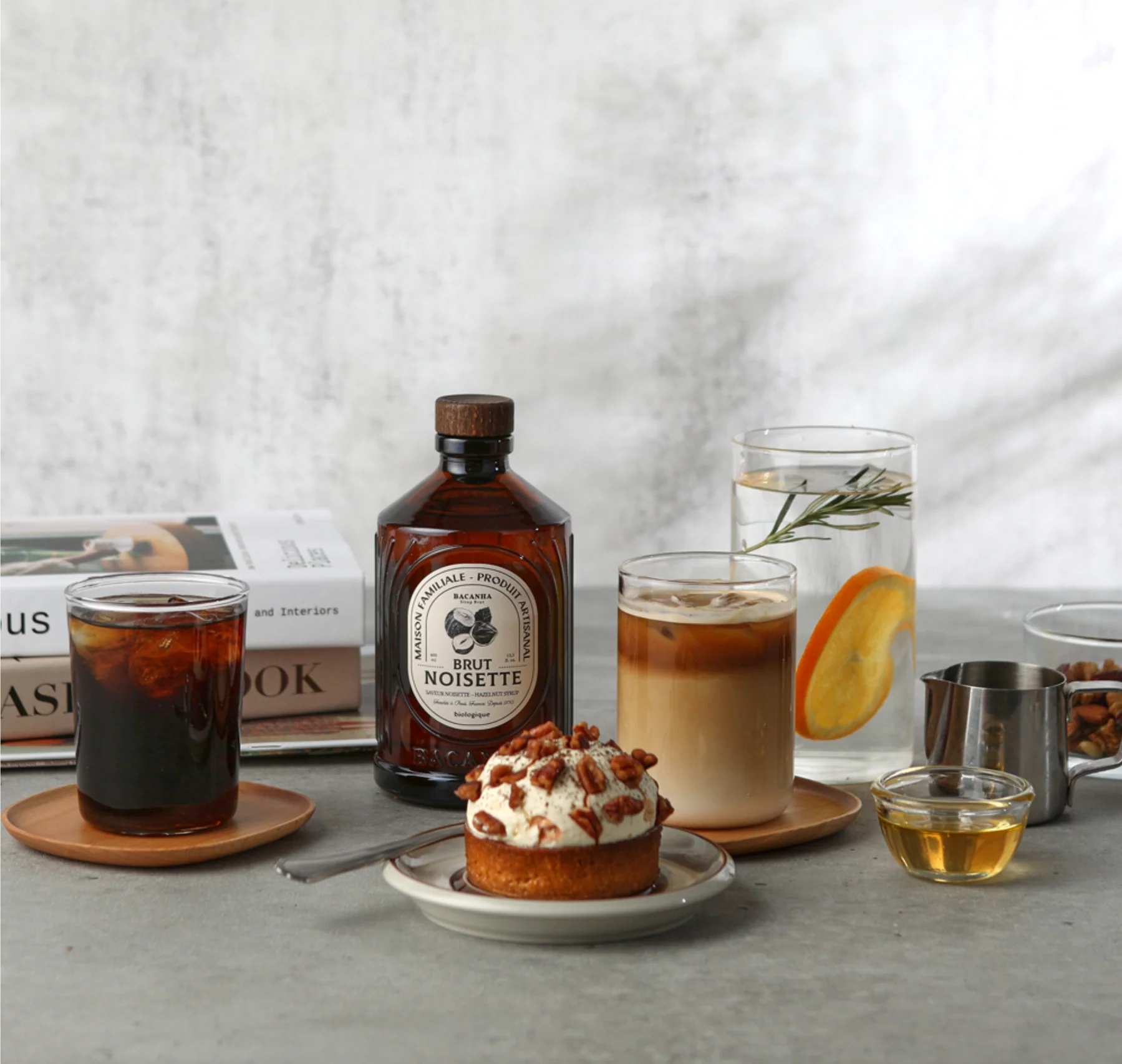 Bacanha Brut Noisette står på et bord sammen med iskaffe, latte, dessert med nøtter og et glass vann med sitrus.