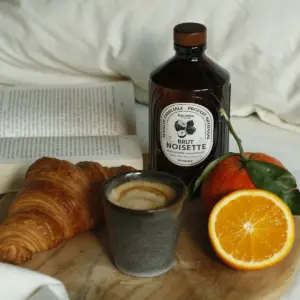 Bacanha Brut Noisette står på et trebord ved siden av croissant, espresso og appelsin i mykt dagslys.