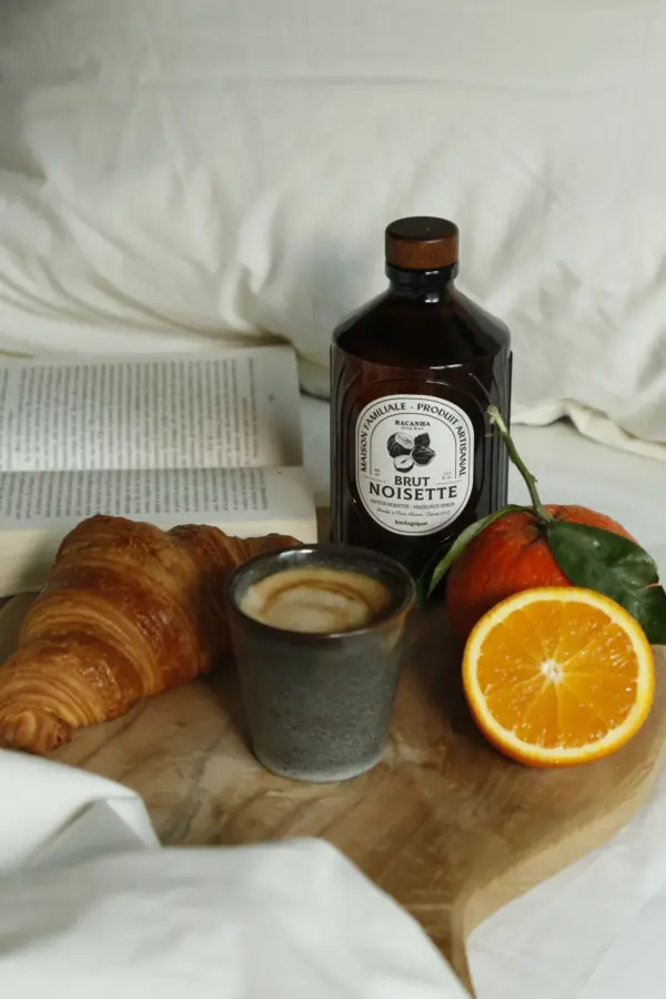 Bacanha Brut Noisette står på et trebord ved siden av croissant, espresso og appelsin i mykt dagslys.