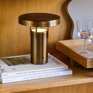 BringMe oppladbar og dimbar bordlampe Mini i Burnished Brass fra Hübsch plassert på en bokhylle, med varmt tent lys.