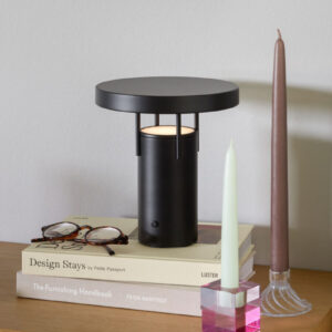 BringMe oppladbar og dimbar bordlampe Mini i Matt Black fra Hübsch plassert på et bord med bøker og dekor, med varmt tent lys.