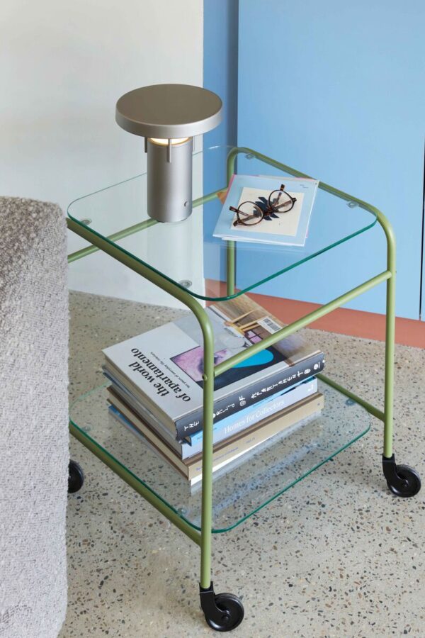 BringMe oppladbar og dimbar bordlampe Mini i Metallic Sand fra Hübsch plassert på et glassbord med bøker, med varmt tent lys.