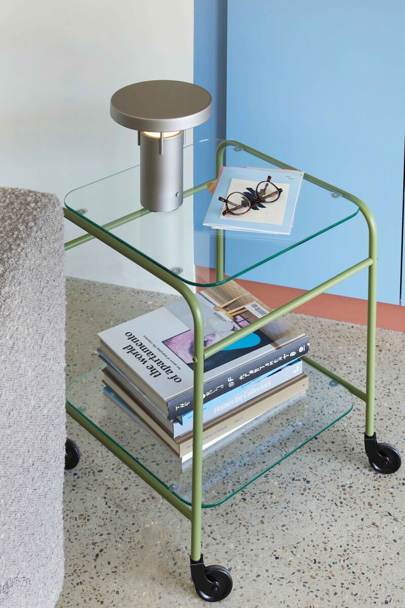 BringMe oppladbar og dimbar bordlampe Mini i Metallic Sand fra Hübsch plassert på et glassbord med bøker, med varmt tent lys.