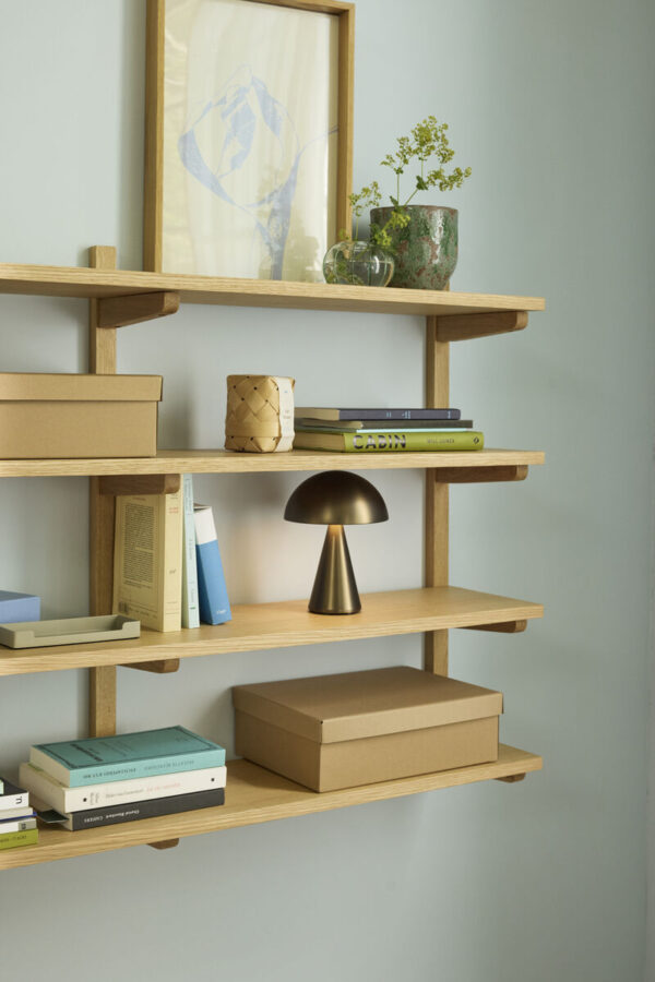 Mush oppladbar og dimbar bordlampe i Burnished Brass fra Hübsch plassert i en hylle, med varmt stemningslys og mykt uttrykk.