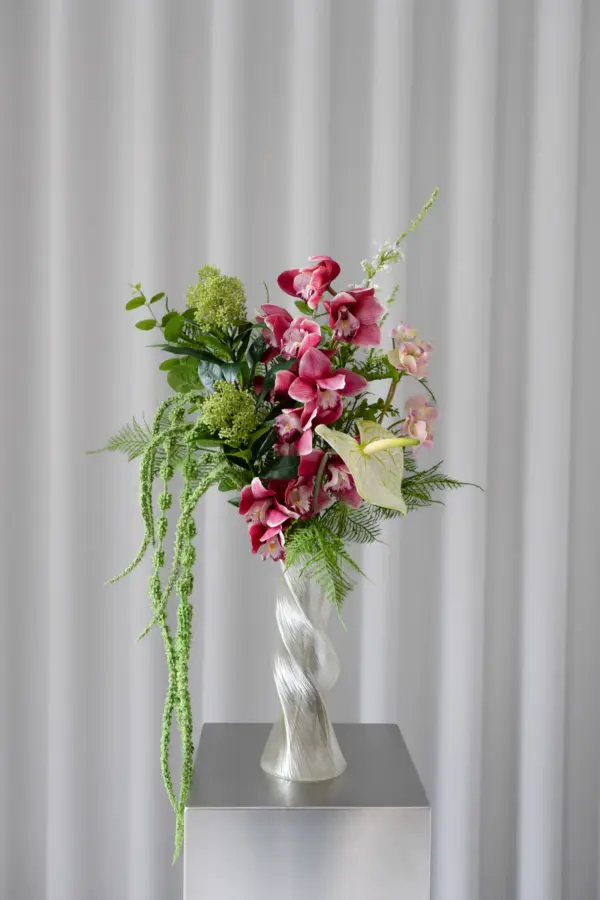 Blush Bloom Bouquet presentert i en elegant, vridd glassvase. De rosa blomstene kombineres med frodige grønne toner og hengende amaranthus som skaper høyde og bevegelse – et sofistikert blikkfang i rommet.